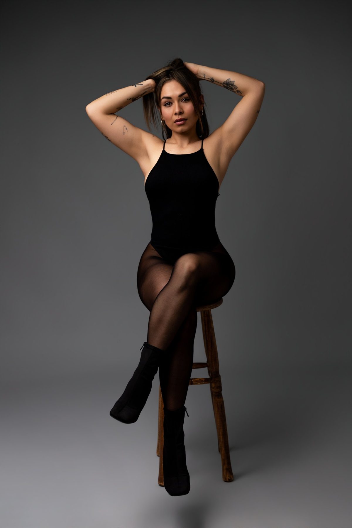 Retrato de estudio de una mujer sentada en un taburete de madera, con las manos en el pelo y mirada directa a cámara