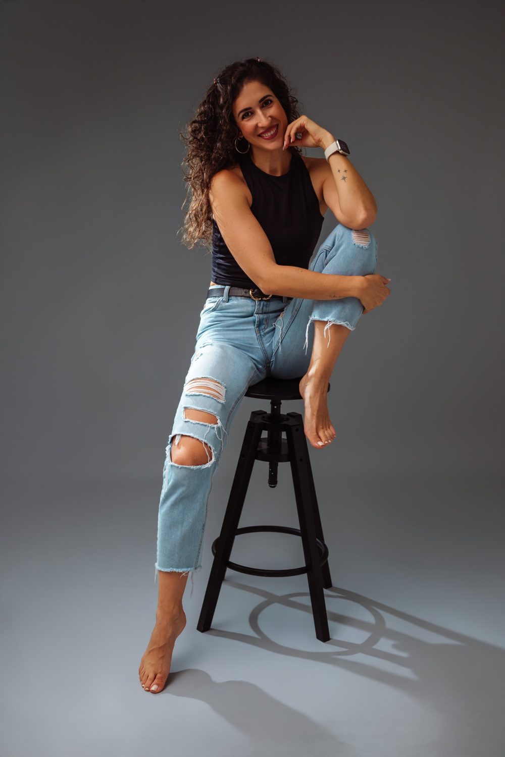 Tali sentada en un taburete del estudio, sonriendo con la mano en la barbilla
